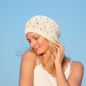 May include: A woman with blonde hair is wearing a white crocheted beanie with a delicate openwork pattern. She is wearing a white tank top and has a soft smile on her face.