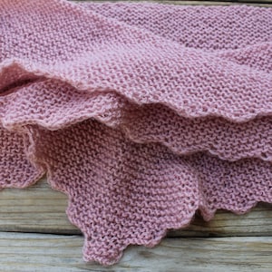 K&ouml;nnte beinhalten: Nahaufnahme eines gestrickten Schals in einem zarten Rosaton. Der Schal hat einen gewellten Rand und ist &uuml;ber einer Holzoberfl&auml;che drapiert. Das Strickmuster ist sichtbar und zeigt die Textur des Garns.