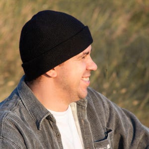 May include: A man wearing a black beanie and a gray denim jacket. He is looking off to the side and smiling.