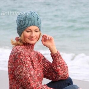 May include: A blue cable-knit beanie with a ribbed brim and the text KnitsMagic printed in white. The beanie is paired with a long-sleeved, high-neck sweater featuring a multi-colored knit pattern in shades of red, grey, and black, and blue jeans.