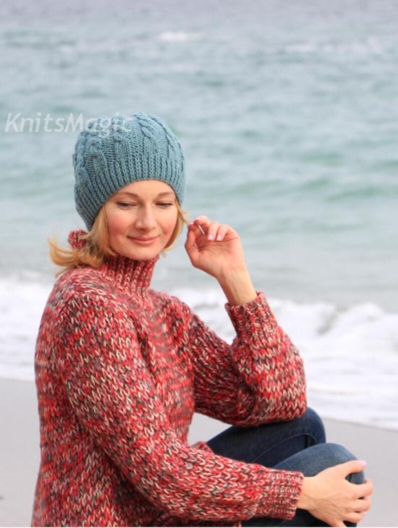 May include: A blue cable-knit beanie with a ribbed brim and the text KnitsMagic printed in white. The beanie is paired with a long-sleeved, high-neck sweater featuring a multi-colored knit pattern in shades of red, grey, and black, and blue jeans.
