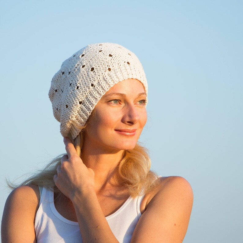 May include: A woman with blonde hair is wearing a white crocheted beanie hat. She is looking off to the side and smiling.