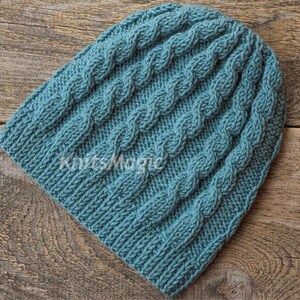 May include: A teal blue knitted beanie featuring a vertical cable knit pattern throughout the fabric. The word KnitsMagic is printed in white across the center of the hat. The beanie has a ribbed edge at the bottom and a rounded top crown.