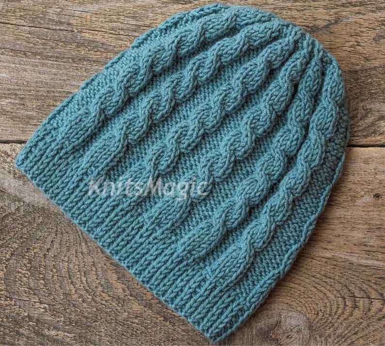 May include: A teal blue knitted beanie featuring a vertical cable knit pattern throughout the fabric. The word KnitsMagic is printed in white across the center of the hat. The beanie has a ribbed edge at the bottom and a rounded top crown.