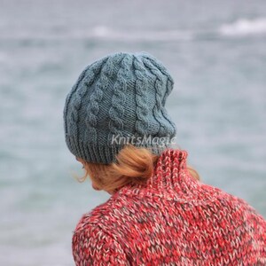 May include: A blue-grey cable knit beanie with a ribbed brim and the text KnitsMagic printed in white. The hat features a vertical braided pattern throughout the crown and is worn over light brown hair, paired with a red and grey textured sweater.