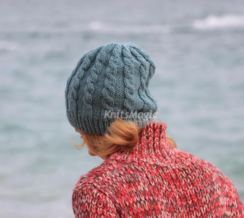 May include: A blue-grey cable knit beanie with a ribbed brim and the text KnitsMagic printed in white. The hat features a vertical braided pattern throughout the crown and is worn over light brown hair, paired with a red and grey textured sweater.