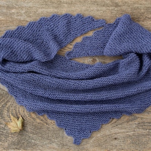 K&ouml;nnte beinhalten: Ein blauer Strickschal mit gewelltem Rand. Der Schal ist aus weichem, strukturiertem Garn gefertigt und liegt auf einer Holzoberfl&auml;che. Ein getrocknetes Blatt liegt in der N&auml;he des Schals.