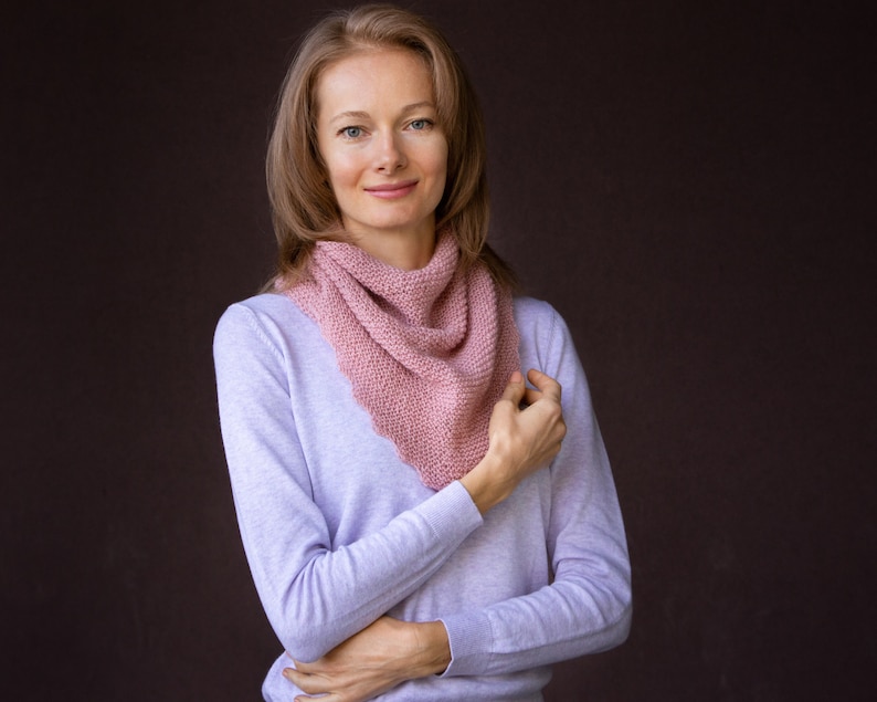 K&ouml;nnte beinhalten: Ein hellrosa gestrickter Schal wird um den Hals getragen. Der Schal ist aus einem weichen, strukturierten Garn gefertigt. Die Person tr&auml;gt ein hellviolettes Langarmshirt. Der Hintergrund ist einfarbig dunkelbraun.