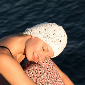 May include: A woman wearing a white crocheted hat with a floral pattern, sitting with her head down and eyes closed. She is wearing a black tank top and a floral skirt.
