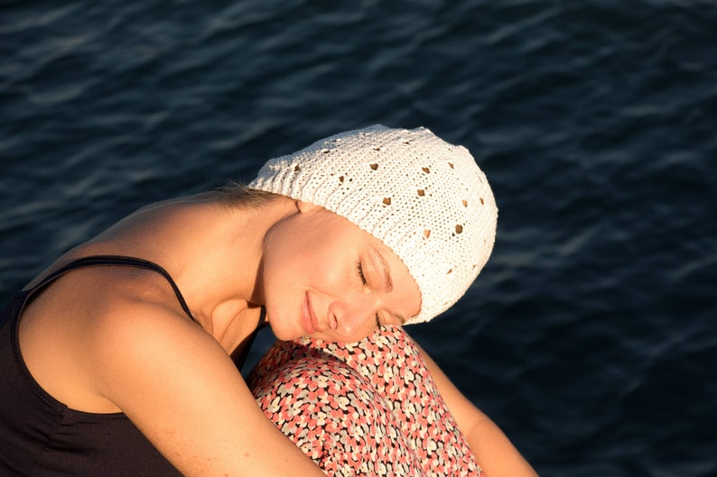 May include: A woman wearing a white crocheted hat with a floral pattern, sitting with her head down and eyes closed. She is wearing a black tank top and a floral skirt.