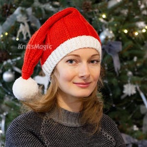 Op de afbeelding: Een felrode gebreide muts met een brede witte geribbelde rand en een witte pompon aan de punt. De muts heeft een verticale ribbelstructuur over het rode gedeelte en wordt op het hoofd gedragen, wat het klassieke kerstman-ontwerp laat zien.