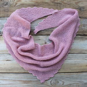 K&ouml;nnte beinhalten: Ein altrosa gestrickter Schal mit gewelltem Rand. Der dreieckige Schal ist auf einer verwitterten Holzoberfl&auml;che drapiert. Das weiche, strukturierte Strickwerk ist ein gem&uuml;tliches Accessoire.