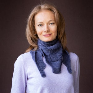 Könnte beinhalten: Eine Frau trägt einen helllila Pullover mit einem blauen Strick Schal um den Hals. Der Schal hat ein geometrisches Muster und ist in einem lockeren Knoten gebunden.