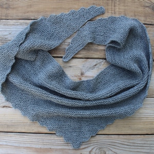 K&ouml;nnte beinhalten: Ein grauer Strickschal mit dreieckiger Form und gewelltem Rand. Der Schal ist aus einem weichen, strukturierten Garn gefertigt und liegt auf einer Holzoberfl&auml;che. Die Gesamt&auml;sthetik ist gem&uuml;tlich und handgemacht.