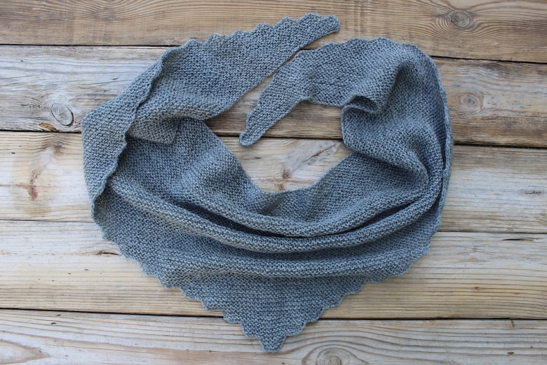 K&ouml;nnte beinhalten: Ein grauer Strickschal mit dreieckiger Form und gewelltem Rand. Der Schal ist aus einem weichen, strukturierten Garn gefertigt und liegt auf einer Holzoberfl&auml;che. Die Gesamt&auml;sthetik ist gem&uuml;tlich und handgemacht.