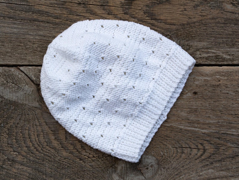 May include: A white crocheted hat with a lacy pattern. The hat is lying on a wooden surface.