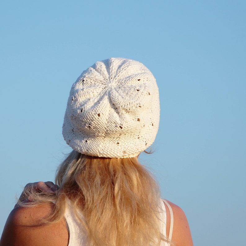 May include: A white crocheted hat with a lacy design. The hat is worn by a person with long blonde hair.