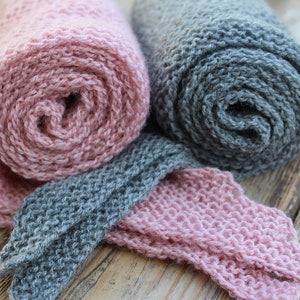 K&ouml;nnte beinhalten: Zwei gerollte Strickschals, einer rosa und einer grau, mit einem Abschnitt der grauen und rosa Schals ausgelegt. Die Schals sind aus einem weichen, strukturierten Garn gefertigt. Der Hintergrund ist eine Holzoberfl&auml;che.