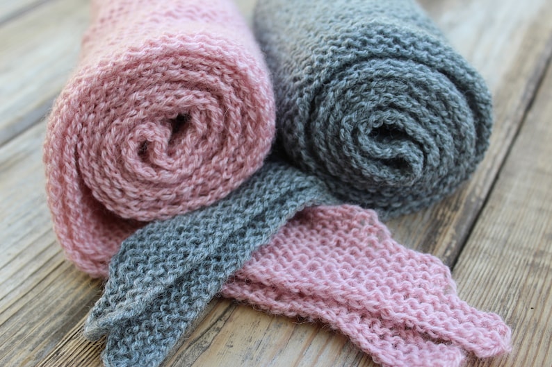 K&ouml;nnte beinhalten: Zwei gerollte Strickschals, einer rosa und einer grau, mit einem Abschnitt der grauen und rosa Schals ausgelegt. Die Schals sind aus einem weichen, strukturierten Garn gefertigt. Der Hintergrund ist eine Holzoberfl&auml;che.