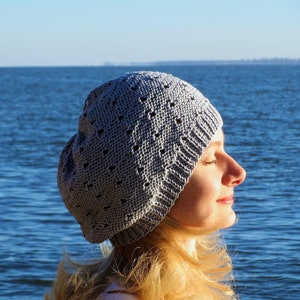 May include: A woman wearing a gray knitted beanie with a lacy pattern, standing in front of a blue body of water.