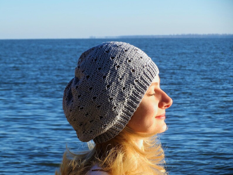 May include: A woman wearing a gray knitted beanie with a lacy pattern, standing in front of a blue body of water.