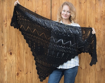 May include: A black crocheted shawl with a delicate lace pattern. The shawl is held by a person in front of a wooden background.