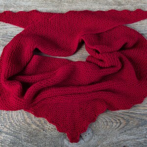 K&ouml;nnte beinhalten: Ein leuchtend roter Strickschal mit einem gewellten Rand. Der Schal ist aus dickem, strukturiertem Garn gefertigt und liegt auf einer Holzoberfl&auml;che. Das Design hat eine einzigartige Form, ideal f&uuml;r W&auml;rme und Stil.