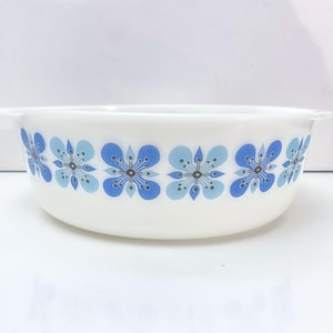 Pode incluir: Tigela de cerâmica branca com um padrão floral azul. A tigela tem formato arredondado com duas pequenas alças em cada lado. O design floral apresenta flores azuis e azul claro com centros azul escuro.