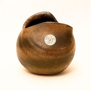 Könnte beinhalten: Eine braune Keramikvase mit einer einzigartigen Form, die einen abgerundeten Körper und eine gebogene Öffnung an der Oberseite aufweist. Die Vase hat eine strukturierte Oberfläche und einen kleinen, kreisförmigen Aufkleber mit Text. Der Farbverlauf reicht von hell- bis dunkelbraun.