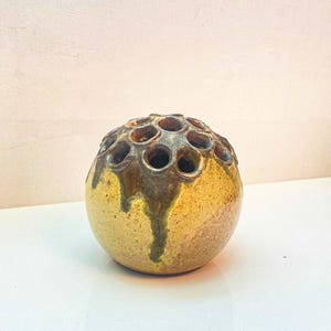 Può includere: Un ranocchio per fiori sferico in ceramica con una glassa maculata, marrone e beige. La parte superiore presenta numerosi fori per la disposizione degli steli. La glassa marrone scuro gocciola lungo i lati. L'oggetto è su una superficie bianca.