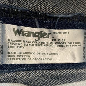May include: Close-up of a Wrangler jeans label. The label is white with black text, including the brand name, size 28 x 32 inches, and care instructions. The fabric is 100% cotton, made in Mexico.