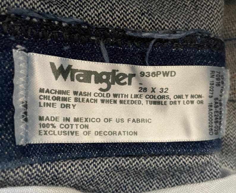 May include: Close-up of a Wrangler jeans label. The label is white with black text, including the brand name, size 28 x 32 inches, and care instructions. The fabric is 100% cotton, made in Mexico.