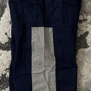 Pu&ograve; includere: Pantaloni sportivi blu navy con una striscia verticale grigio chiaro al centro. La parola "NIKE" &egrave; stampata in verde sulla striscia grigia. Una piccola cerniera dorata &egrave; visibile nella parte superiore.