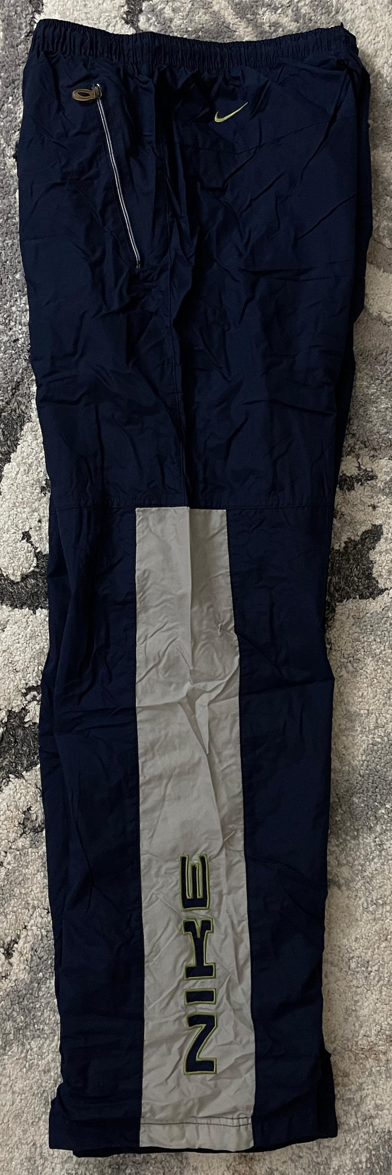 Pu&ograve; includere: Pantaloni sportivi blu navy con una striscia verticale grigio chiaro al centro. La parola "NIKE" &egrave; stampata in verde sulla striscia grigia. Una piccola cerniera dorata &egrave; visibile nella parte superiore.