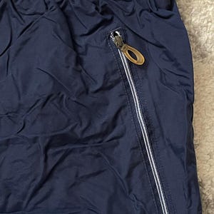Pu&ograve; includere: Primo piano di pantaloni sportivi blu navy. I pantaloni hanno un elastico in vita e una tasca con cerniera argentata con un tirante dorato. Il tessuto ha una leggera lucentezza.