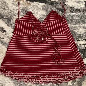 May include: Red and white striped tank top with a V-neck and a decorative lace-up back. The bottom hem features a scalloped edge with white embroidery. The straps are thin and adjustable.