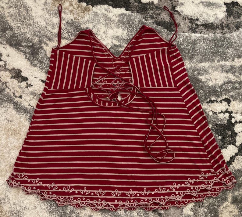 May include: Red and white striped tank top with a V-neck and a decorative lace-up back. The bottom hem features a scalloped edge with white embroidery. The straps are thin and adjustable.