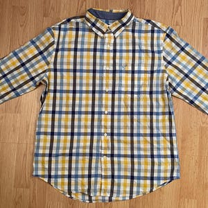 May include: A long-sleeve button-down shirt featuring a plaid pattern in shades of yellow, navy blue, and light blue. The shirt has a standard collar, a front button placket, and a single chest pocket on the left side with a small embroidered logo.