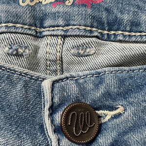Puede incluir: Primer plano de unos vaqueros de mezclilla azules con el nombre de la marca "Wrangler" bordado en hilo blanco y rosa. Se ve un bot&oacute;n marr&oacute;n oscuro con el logotipo "W". Los vaqueros tienen costuras blancas.