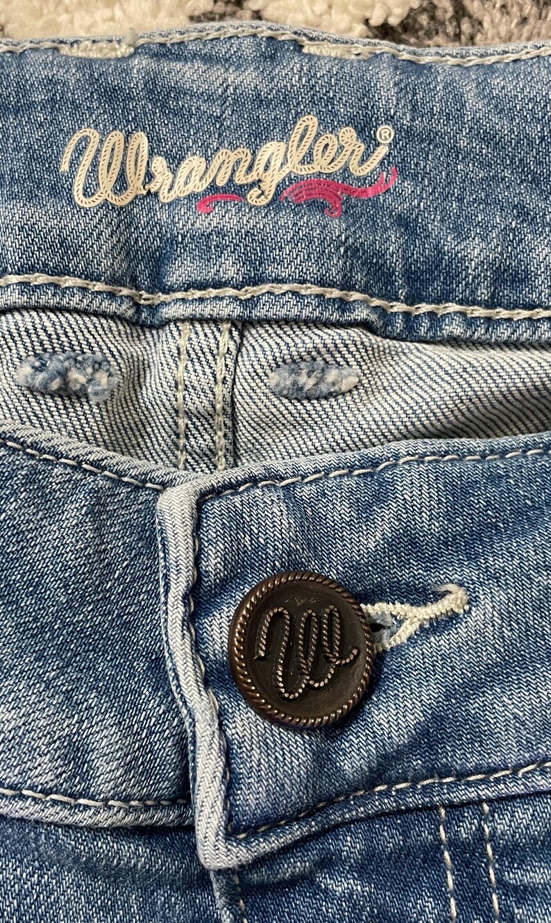 Puede incluir: Primer plano de unos vaqueros de mezclilla azules con el nombre de la marca "Wrangler" bordado en hilo blanco y rosa. Se ve un bot&oacute;n marr&oacute;n oscuro con el logotipo "W". Los vaqueros tienen costuras blancas.