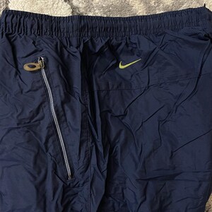 Pu&ograve; includere: Pantaloni sportivi blu navy con elastico in vita e tasca con zip. &Egrave; visibile un piccolo logo Nike swoosh verde neon. I pantaloni hanno una texture lucida simile al nylon.