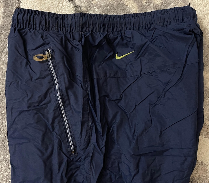 Pu&ograve; includere: Pantaloni sportivi blu navy con elastico in vita e tasca con zip. &Egrave; visibile un piccolo logo Nike swoosh verde neon. I pantaloni hanno una texture lucida simile al nylon.