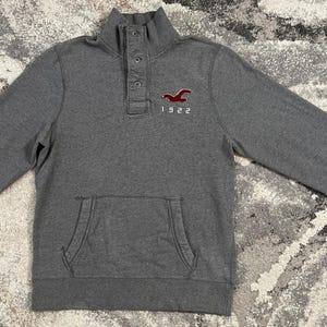 May include: A charcoal gray long-sleeve pullover sweatshirt featuring a mock neck with a button-up placket. The chest displays a red embroidered bird logo above the text 1922. It includes a front kangaroo pocket and ribbed sleeve cuffs.