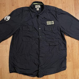 May include: A navy blue long-sleeve button-down shirt featuring two chest pockets, a collar with lime green interior trim, a circular patch on the right sleeve, and a rectangular patch on the left chest pocket that reads North Dakota.