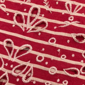 May include: Close-up of red and white striped fabric with white embroidered floral and leaf patterns. The fabric has a textured appearance with a combination of solid and openwork designs. The embroidery includes various shapes, such as circles and teardrop-like forms.