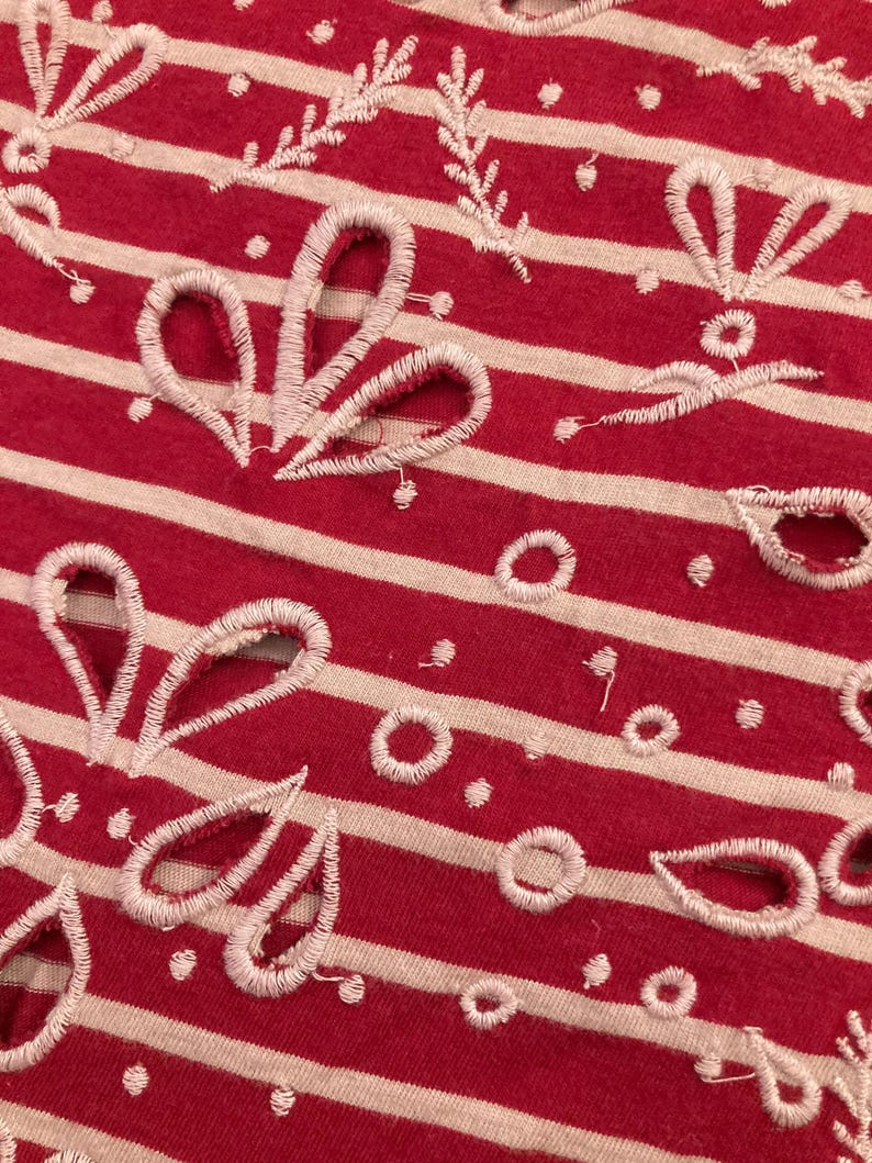 May include: Close-up of red and white striped fabric with white embroidered floral and leaf patterns. The fabric has a textured appearance with a combination of solid and openwork designs. The embroidery includes various shapes, such as circles and teardrop-like forms.