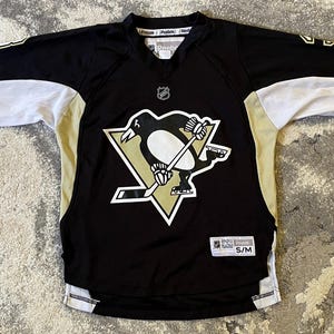 May include: A black, white, and gold Reebok hockey jersey featuring a Pittsburgh Penguins logo on the chest. The jersey has long sleeves with white and gold accents, a V-neck collar, and a white tag at the bottom hem labeled S/M with NHL branding.
