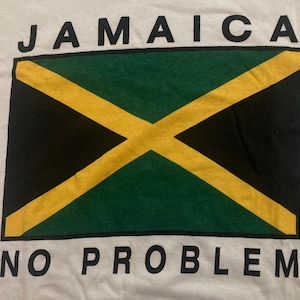 以下が含まれることがあります： ジャマイカの国旗のデザインが施された白いTシャツ。国旗は、緑と黒の背景に黄色の十字が描かれた長方形です。「JAMAICA」と「NO PROBLEM」の文字が、国旗の上と下に黒いブロック体で印刷されています。