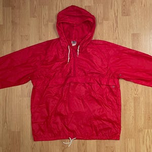 May include: A bright red pullover windbreaker jacket featuring a hood with white drawstring cords, long sleeves, and a front kangaroo pocket. The jacket includes a partial front zipper closure and an adjustable white drawstring at the bottom hem.