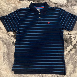 May include: Navy blue polo shirt with horizontal light blue stripes. Features a collared neckline, three buttons, and a small red logo on the chest. The shirt has short sleeves and a classic fit.